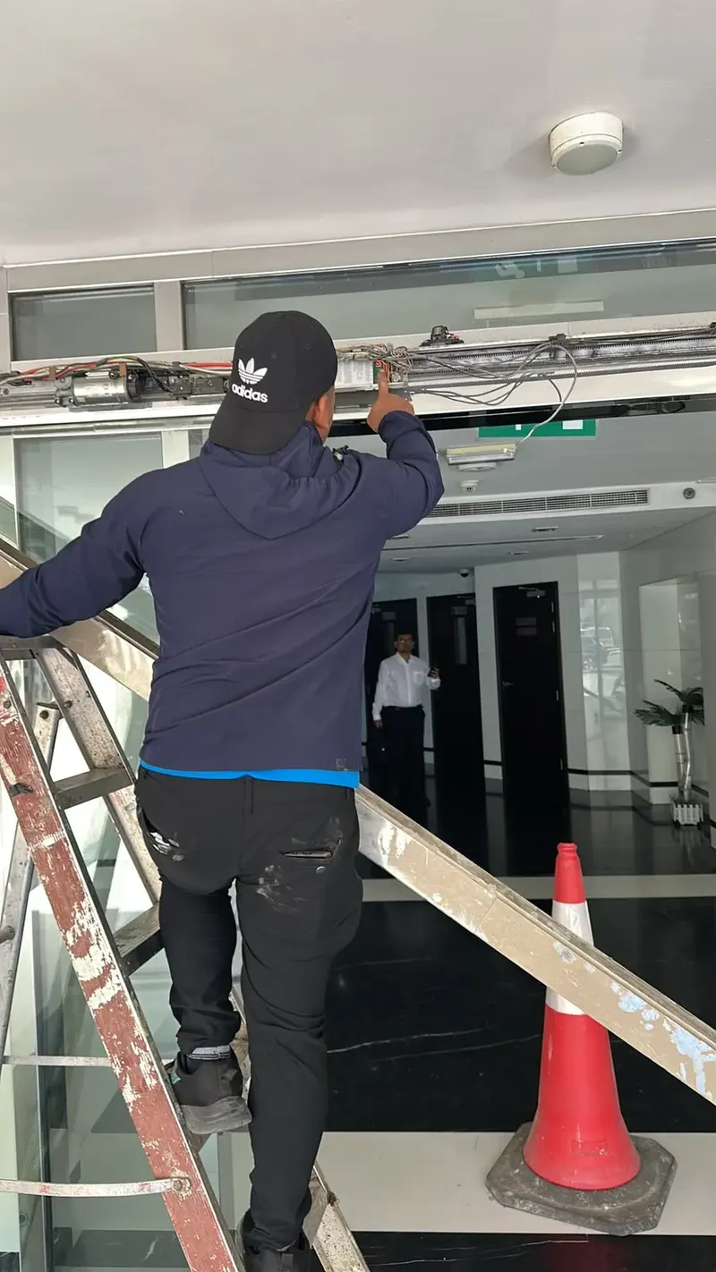 Technician on a ladder checking the open header of an automatic glass door in a lobby in Deira, Dubai