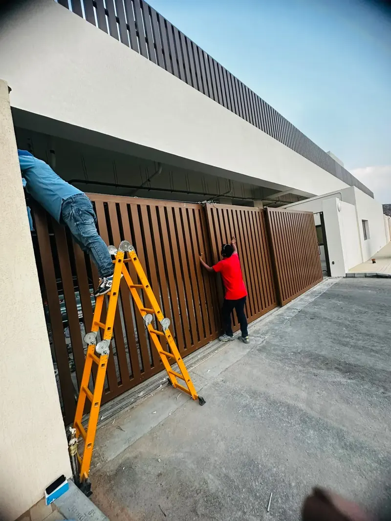 Two technicians repairing a brown three leaf telescopic sliding gate with a folding ladder in Hatta, UAE